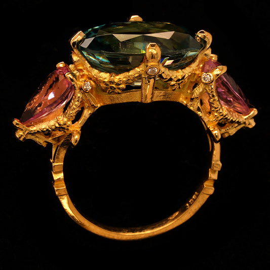An 18ct yellow gold ring featuring a central aqua blue Zircon, flanked by two pink pear cut Tourmalines, with a band detailed with intricate scrollwork and tiny diamonds set throughout.