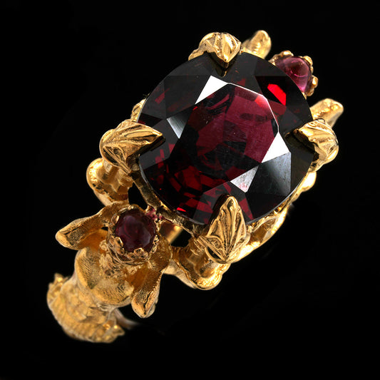 A gold ring featuring a large, faceted garnet central gemstone and smaller matching red accent stones, set against a solid black background.