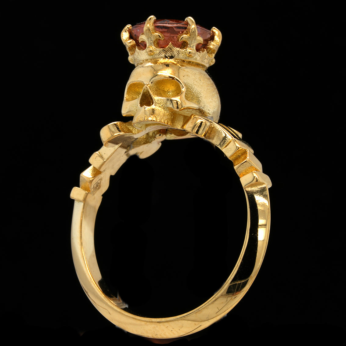 A gold ring featuring a skull topped with a crown and a pink tourmaline gemstone, set against a solid black background.