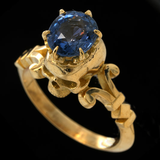 A yellow gold ring featuring an oval-cut blue sapphire gemstone in a decorative skull-like setting with sculpted details on the band, shown against a solid black background.