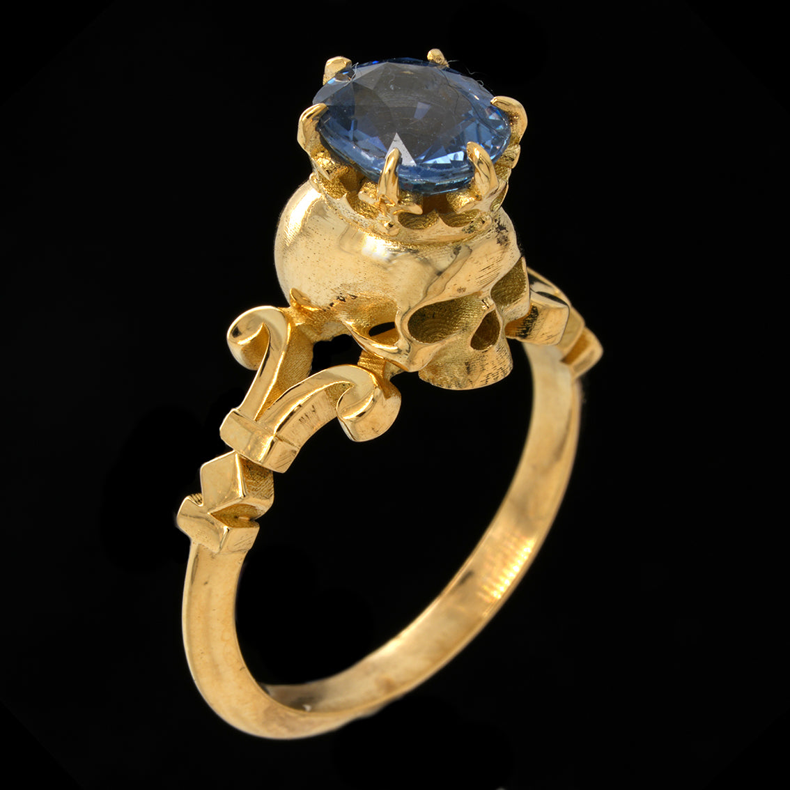 A gold ring featuring a skull motif with an oval blue sapphire on top, set against a solid black background.