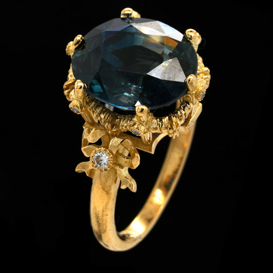 A yellow gold ring featuring a large oval-cut dark blue gemstone in a detailed floral setting, accented with small clear stones on the band, set against a solid black background.