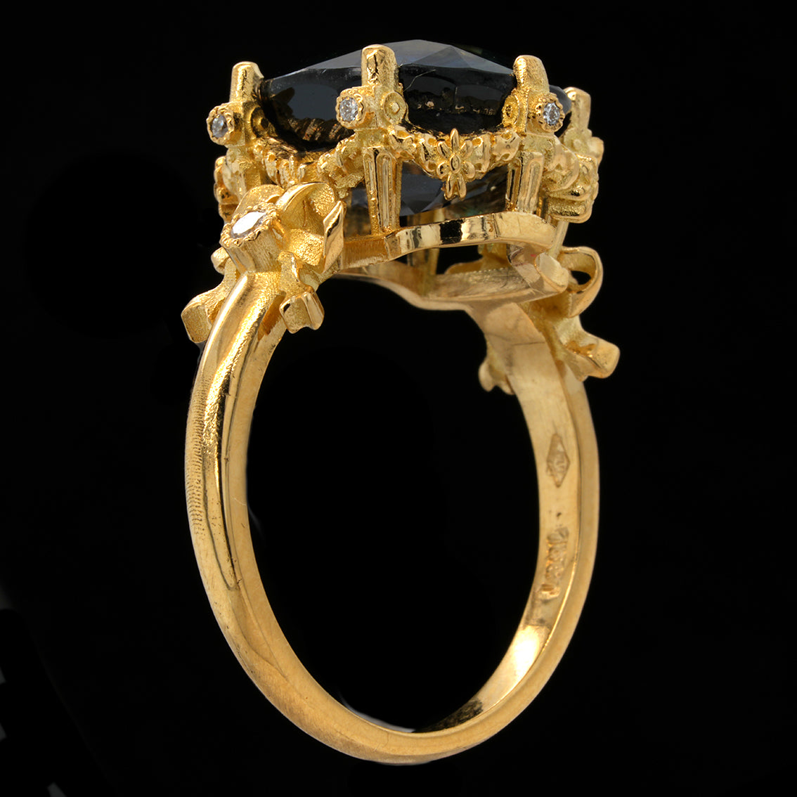 A gold ring featuring a large black oval gemstone in a highly detailed, ornate crown-like setting with small clear accent stones, shown against a solid black background.