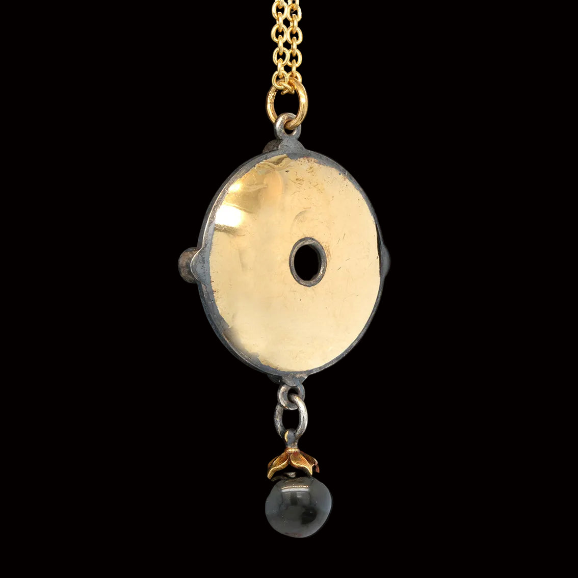 A gold and silver pendant necklace featuring a circular disc with a central hole, suspended from a gold chain with a black pearl dangling below, set against a solid black background.