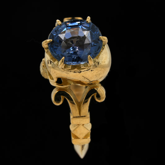 A gold ring featuring a  faceted blue sapphire gemstone held in a claw setting, mounted on an ornate gold skull, shown against a solid black background.