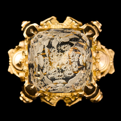 An ornate gold ring featuring a citrine, reverse intaglio cut with intricate floral engravings, set against a solid black background.