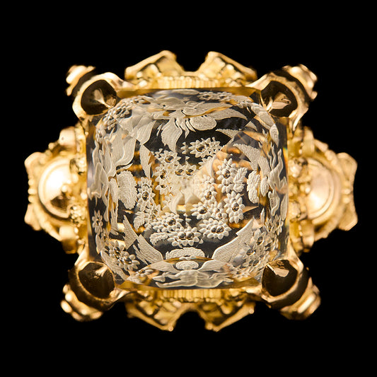 An ornate gold ring featuring a citrine, reverse intaglio cut with intricate floral engravings, set against a solid black background.