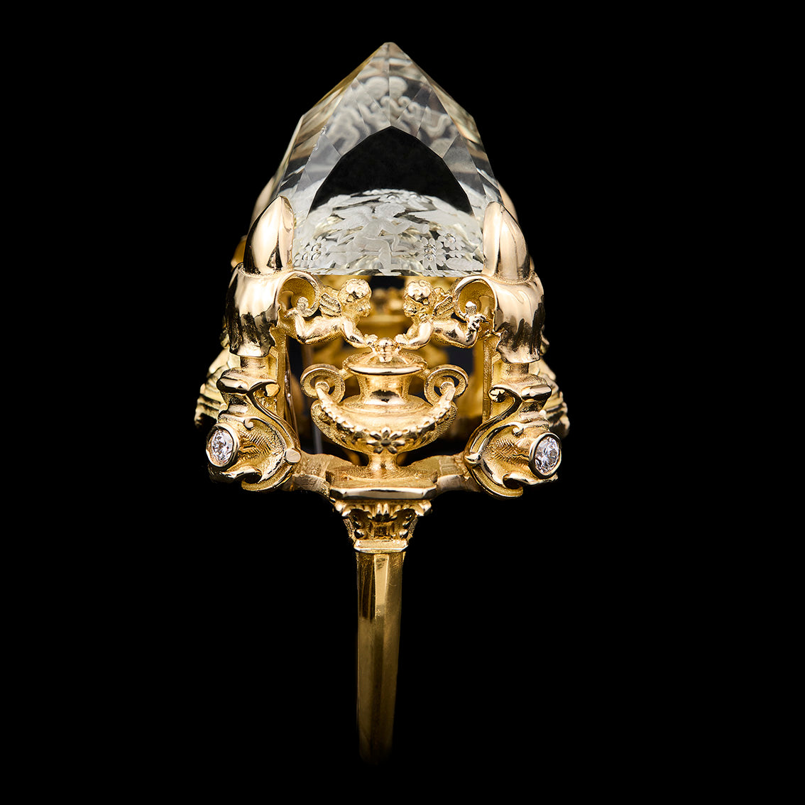 An ornate yellow gold ring featuring a large, faceted clear gemstone on top of a highly detailed sculpted gold head with small accent stones, set against a solid black background.