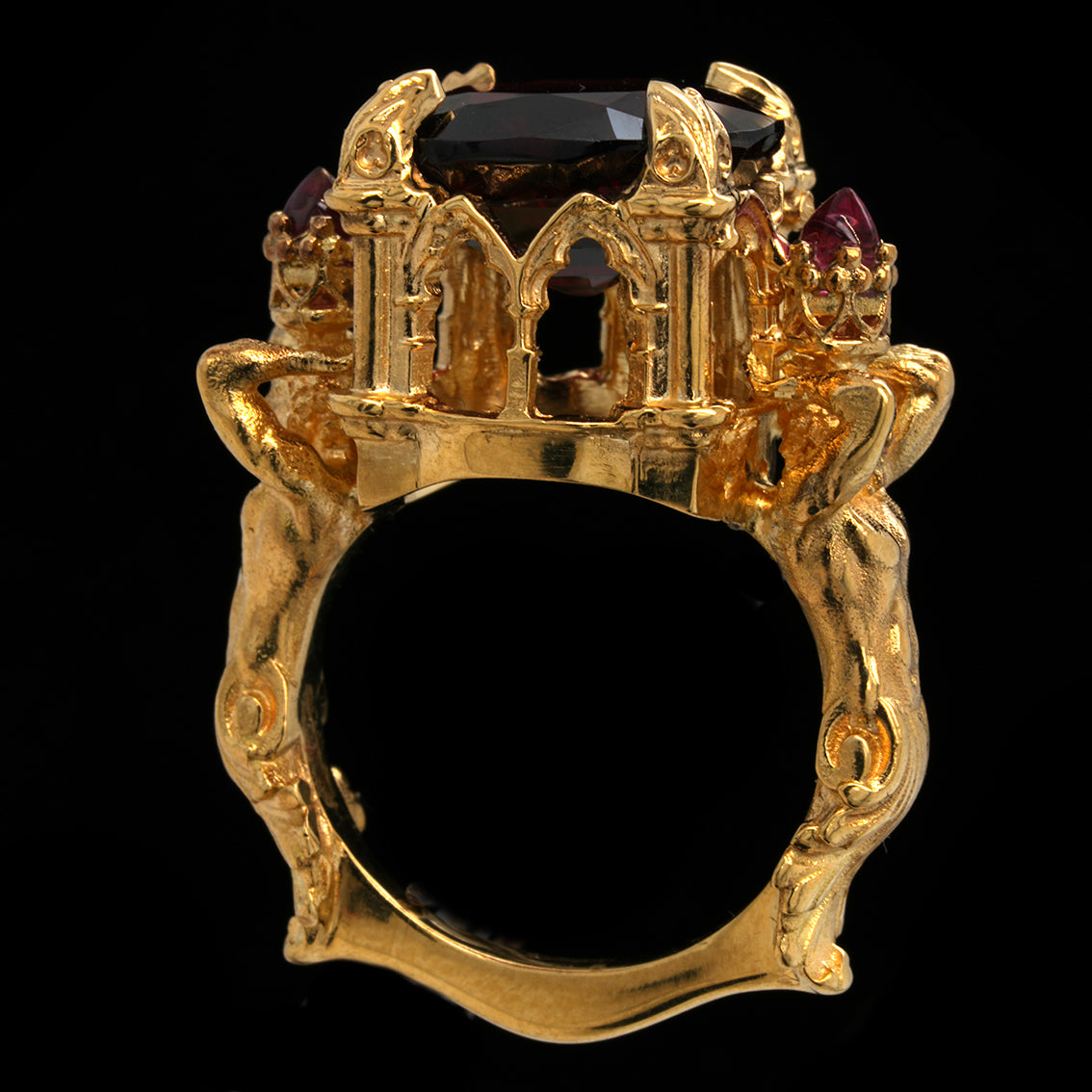 An ornate gold ring featuring a castle-like top setting with a large garnet gemstone and smaller dark red gemstones, shown against a solid black background.