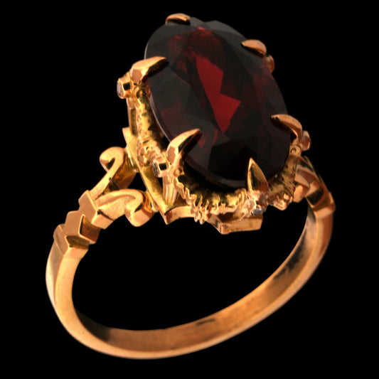 A gold ring featuring a large, faceted garnet gemstone in a claw setting with ornate, floral details on the band, shown against a solid black background.