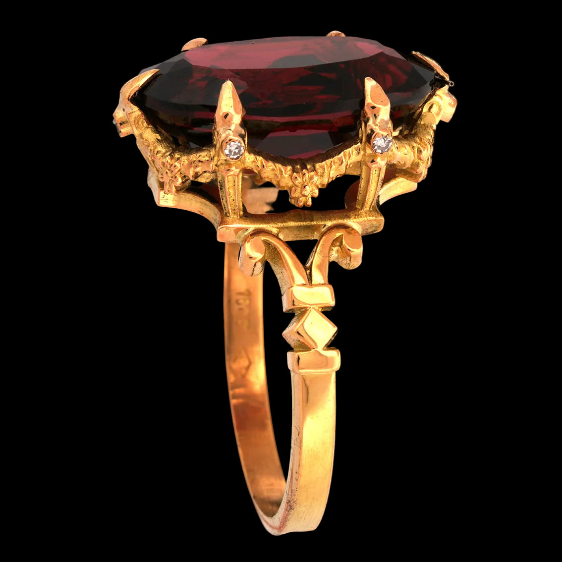 A gold ring featuring a large oval dark red garnet gemstone in a decorative crown-like setting with small diamond accent stones, shown against a solid black background.