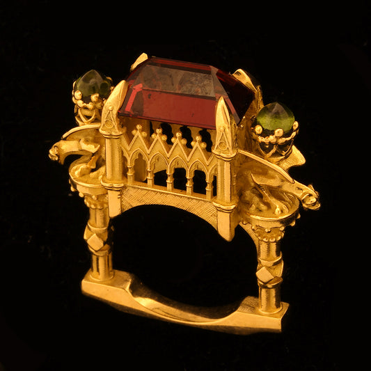 A yellow gold ring with an emerald cut garnet and additional peridot stones, featuring a detailed, cathedral-like structure with gargoyles on the sides.