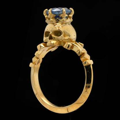 A gold ring featuring a skull-shaped setting topped with a small gold crown and a blue sapphire gemstone, shown against a solid black background.