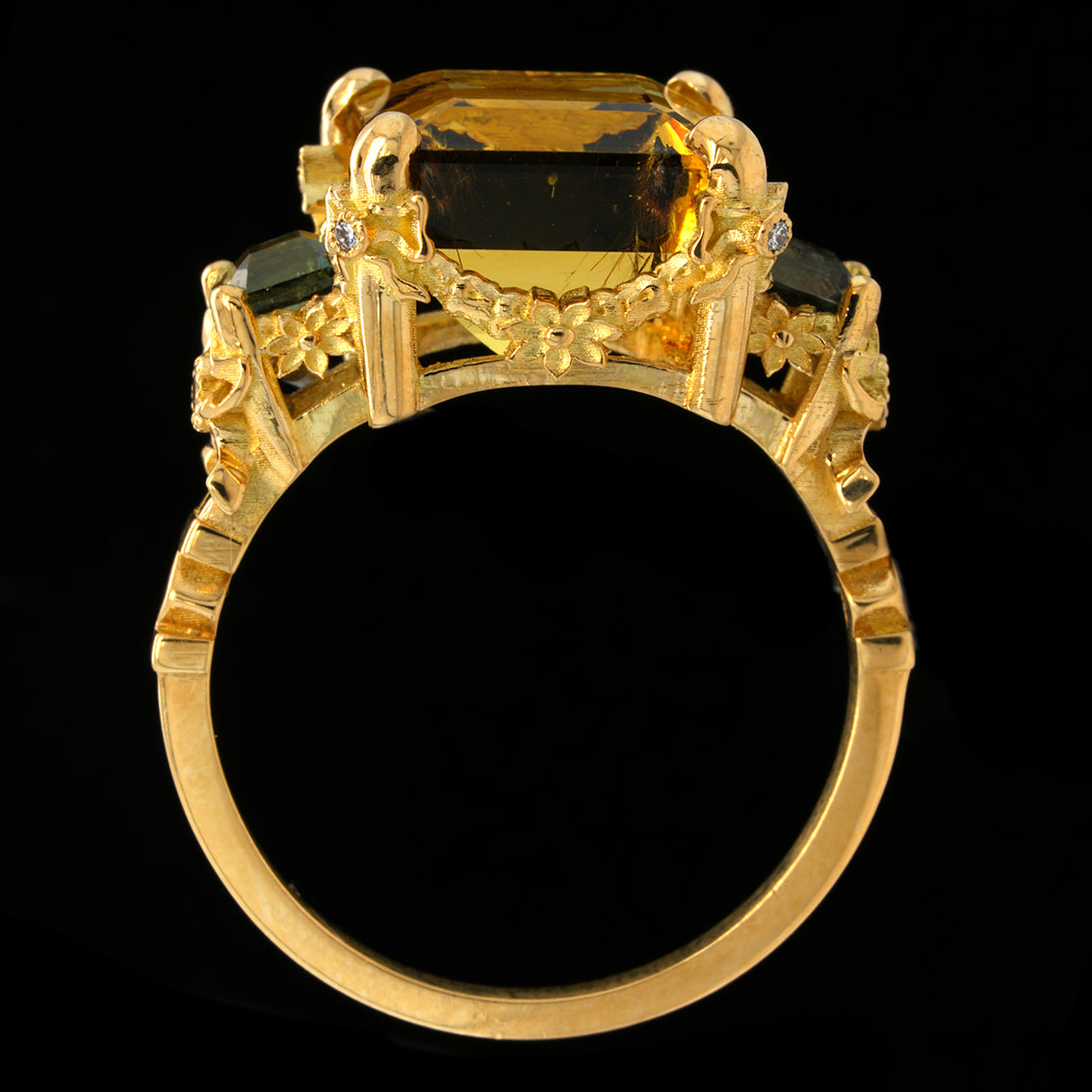 A yellow gold ring featuring a large rectangular yellow gemstone in a decorative crown-like setting with smaller dark green and clear accent stones, shown against a solid black background.