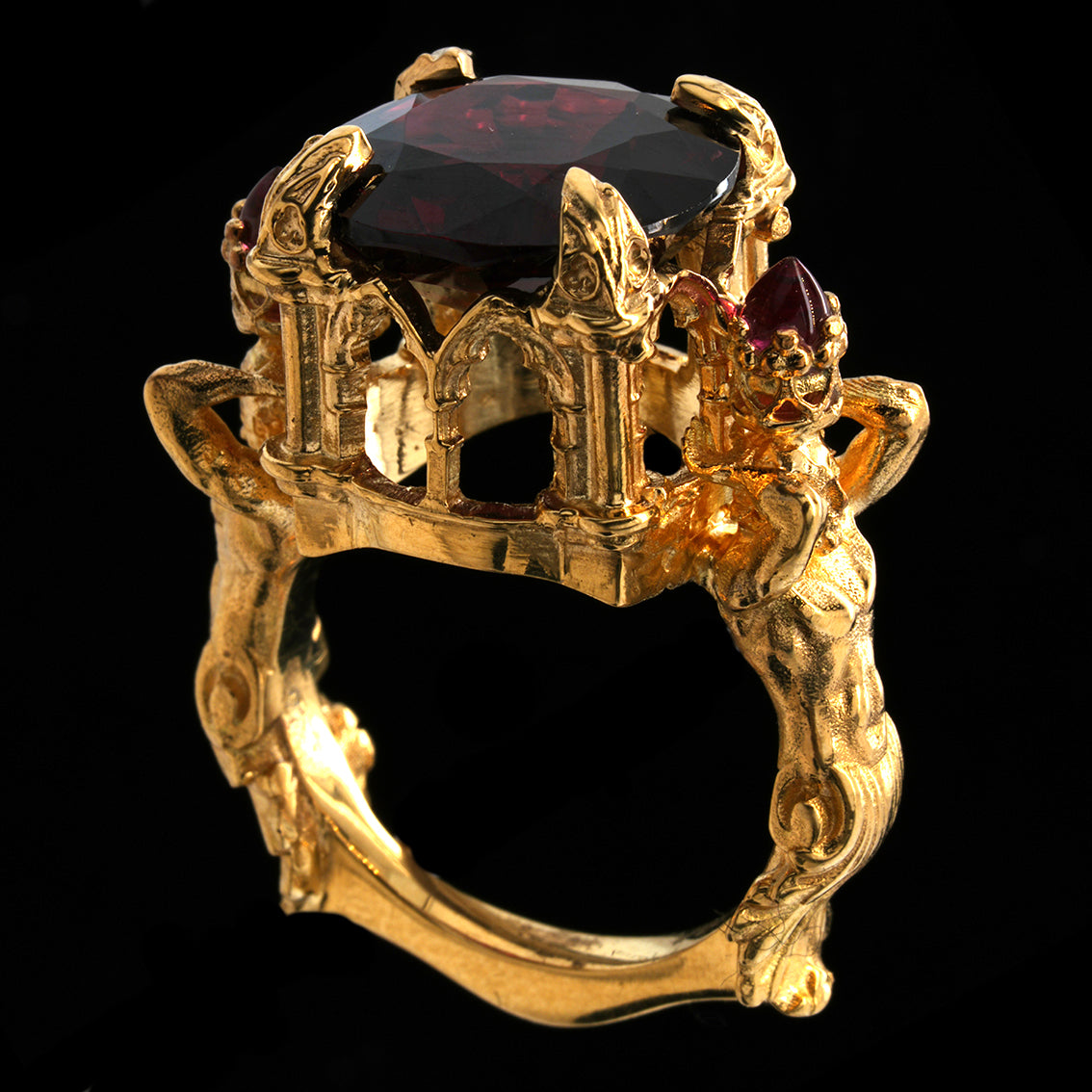 A gold ring featuring a gothic castle crown design with a large garnet on top and smaller red gemstones on the side, set against a solid black background.