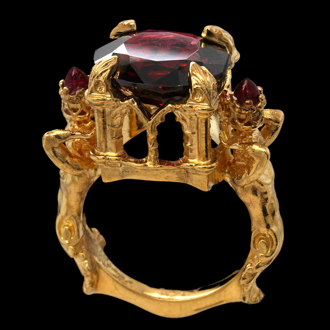 An ornate gold ring featuring a large, faceted garnet gemstone at the center, flanked by smaller red gemstones and decorative sculpted details on the shoulders, set against a solid black background.