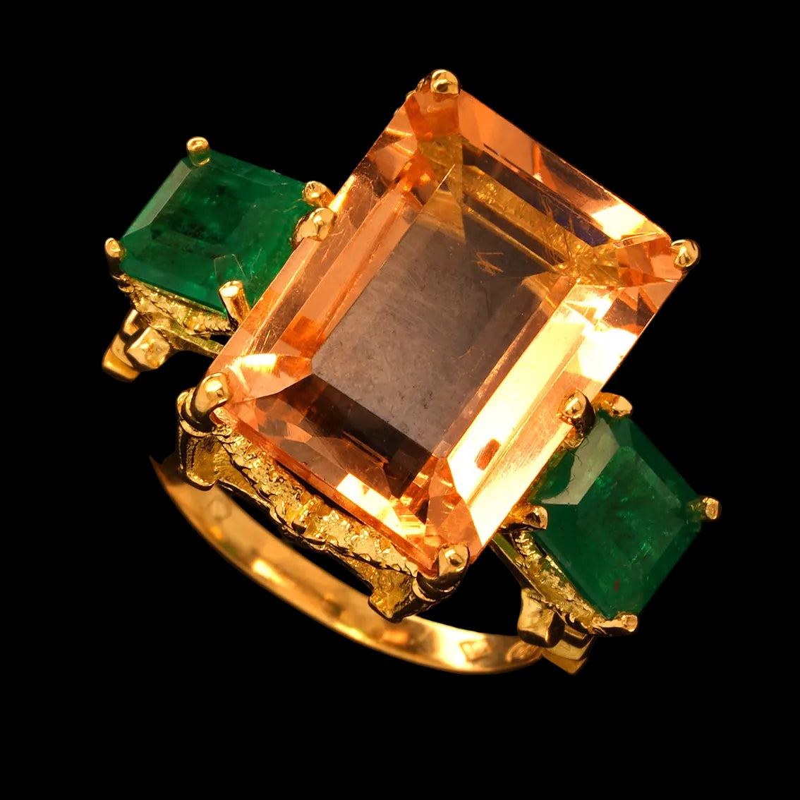 A yellow gold ring featuring a large rectangular emerald-cut morganite center stone, flanked by two rectangular emerald gemstones on the shoulders, set against a solid black background.