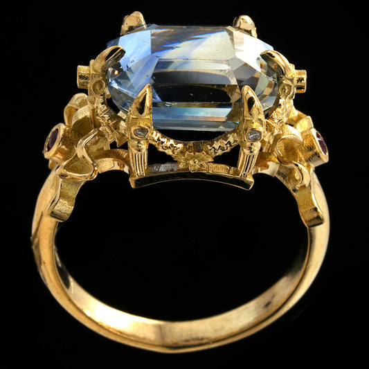A yellow gold ring featuring a large, faceted light blue rectangular gemstone in a crown-like setting with small accent stones, shown against a solid black background.