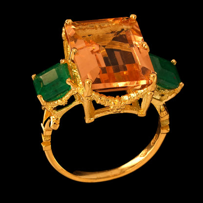 A yellow gold ring featuring a large rectangular peach morganite center stone and two emerald-cut emerald side stones, set against a solid black background.