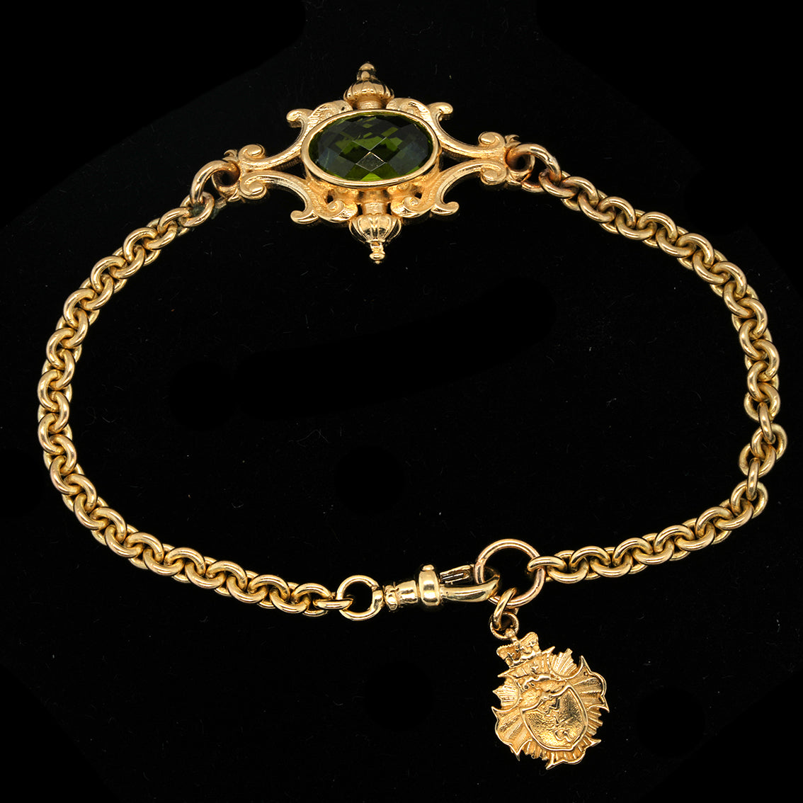 A  baroque style gold chain bracelet with an ornate decorative clasp featuring a peridot oval stone, displayed on a black background.