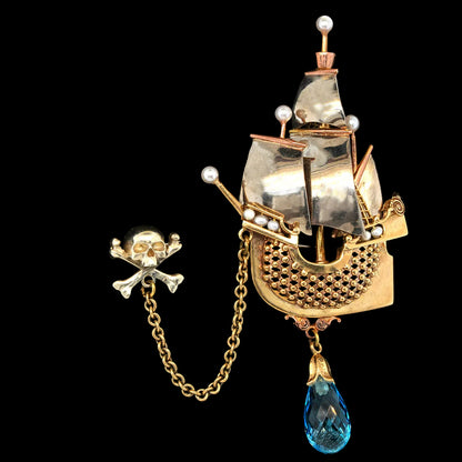 A pirate ship brooch and matching skull-and-crossbones pin on a black background. The ship brooch is gold and silver-tone with white pearl-like accents and a dangling blue teardrop crystal.