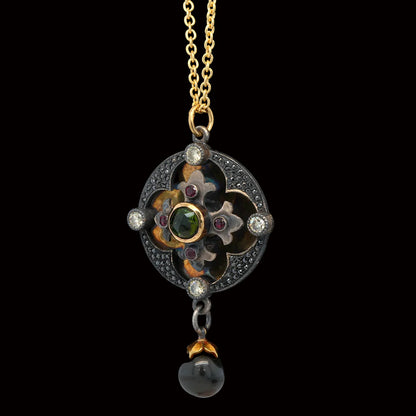 An antique-style pendant necklace featuring a gold chain, a circular floral clover pendant with dark enamel, iridescent green and brown accents, small clear stones, and a dangling dark pearl bead at the bottom, displayed on a black velvet stand.