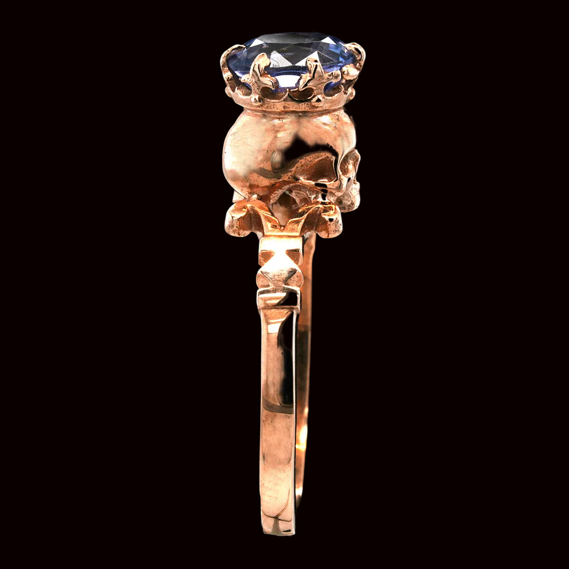 A rose gold skull ring with a crown-like setting and a faceted blue gemstone on top, shown against a solid black background.