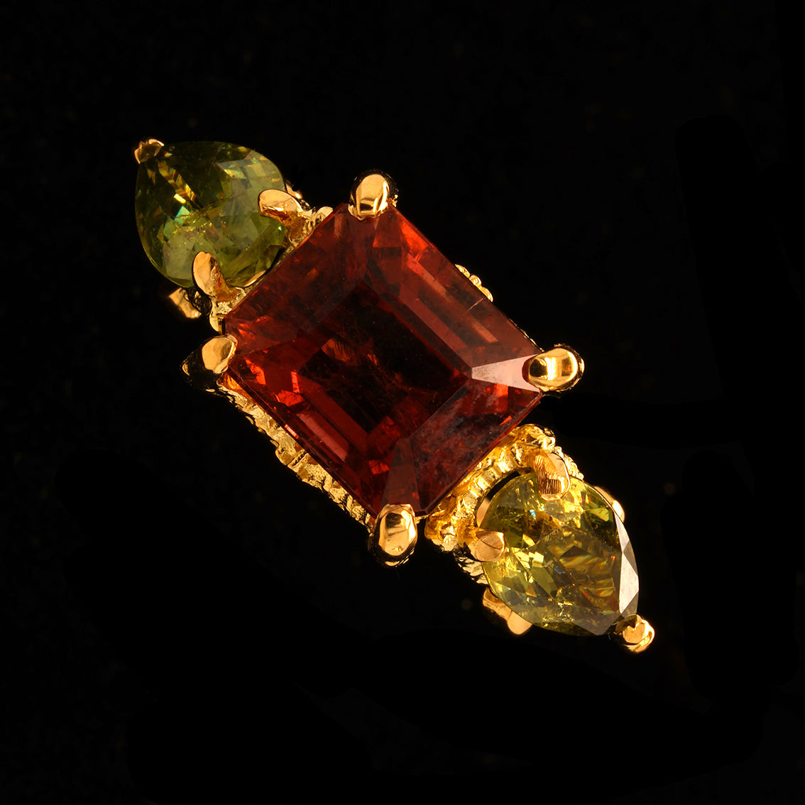A gold ring featuring a large rectangular rubelite tourmaline in the center, flanked by two pear-cut demantoid garnet on either side, set against a solid black background.