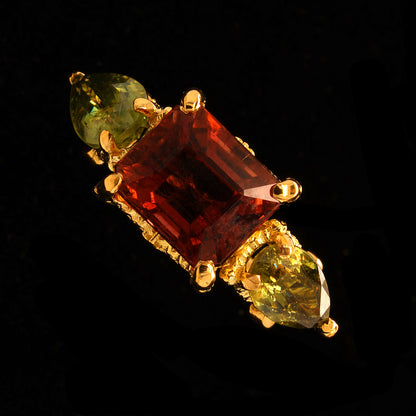 A gold ring featuring a large rectangular rubelite tourmaline in the center, flanked by two pear-cut demantoid garnet on either side, set against a solid black background.
