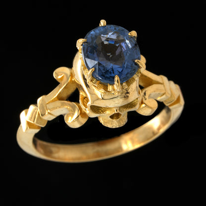 A yellow gold ring featuring an oval-cut blue sapphire in a decorative claw setting with ornate scrollwork on the band, shown against a solid black background.
