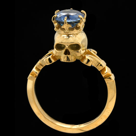 A gold ring featuring a skull at the center with a crown topped by a faceted blue sapphire gemstone, set against a solid black background.