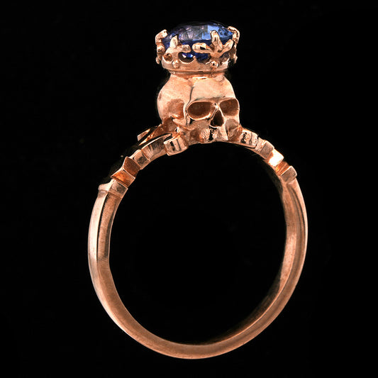 A skull ring in rose gold with a crown-like setting and an oval blue sapphire, shown against a solid black background.