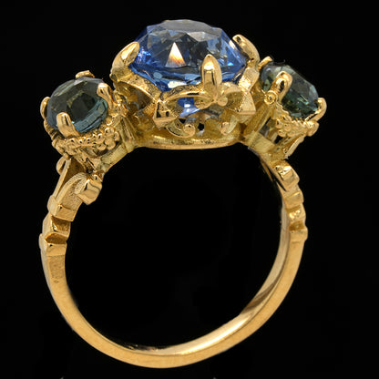 A yellow gold ring featuring a large central faceted blue sapphire and two smaller blue-green teal sapphires on either side, set against a solid black background.