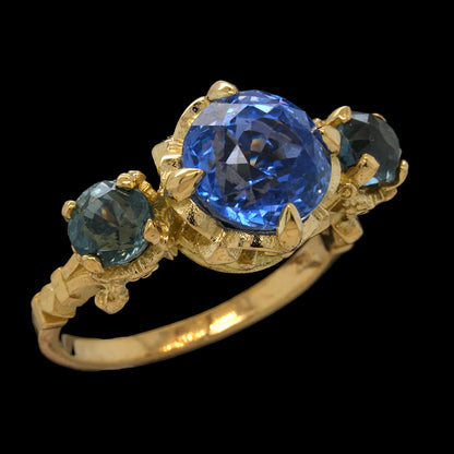 A yellow gold ring featuring a large round-cut blue sapphire center stone and two smaller oval-cut blue sapphires on either side, set against a solid black background.
