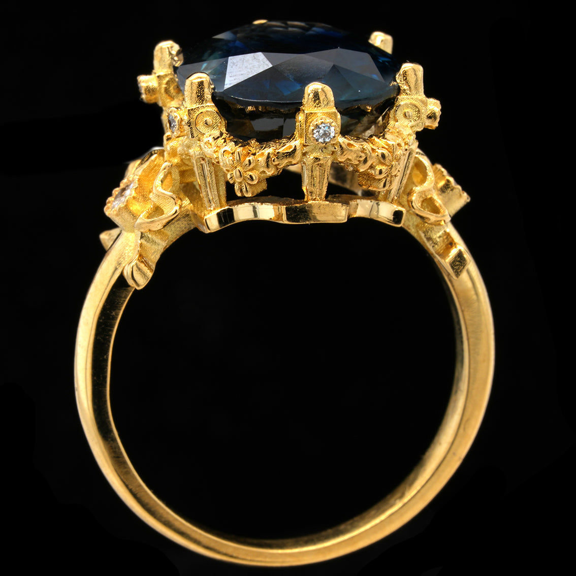 A yellow gold ring featuring a large, faceted teal sapphire in a detailed crown-like setting with small accent diamonds, shown against a solid black background.