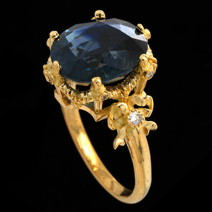 A yellow gold ring featuring a large oval-cut dark blue sapphire center stone, accented with small clear stones in floral details on the shoulders, set against a solid black background.