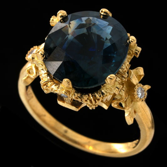 A yellow gold ring featuring a large oval-cut teal sapphire in a detailed floral setting, accented with small diamonds on the band, set against a solid black background.