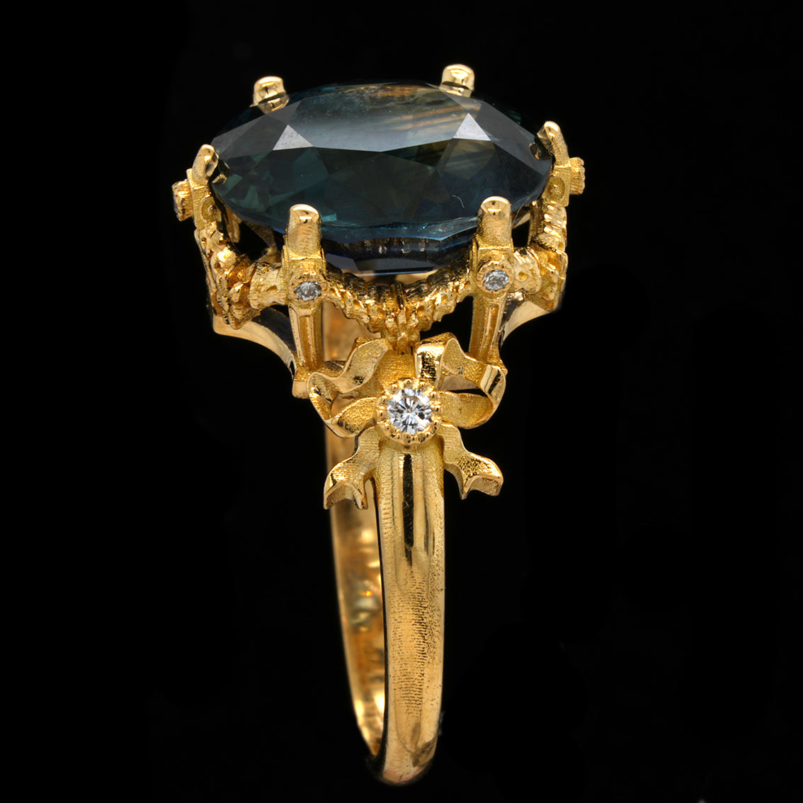 A yellow gold ring featuring a large teal sapphire in a claw setting, accented with small round diamonds on the decorative shoulders and band, set against a solid black background.