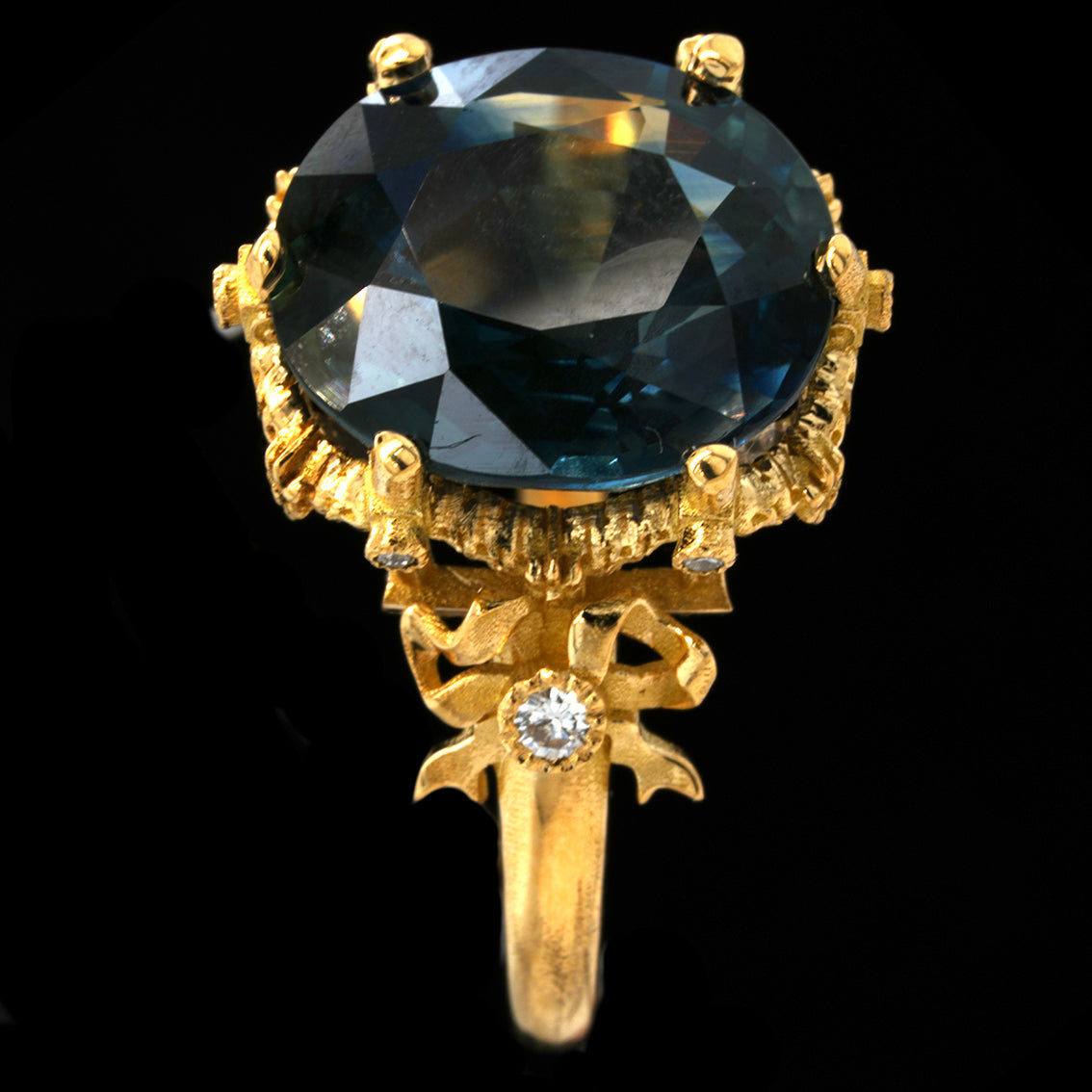 A yellow gold ring featuring a large oval-cut teal sapphire in a detailed claw setting, with a decorative bow motif on the band accented by a small diamond, shown against a solid black background.