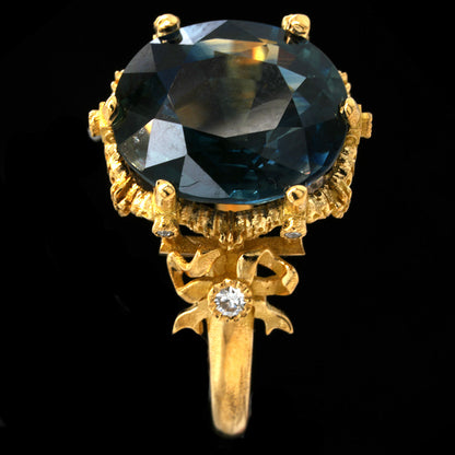 A yellow gold ring featuring a large oval-cut teal sapphire in a detailed claw setting, with a decorative bow motif on the band accented by a small diamond, shown against a solid black background.