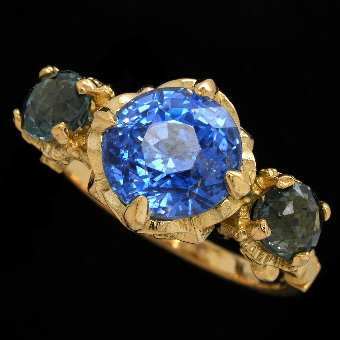 A yellow gold ring featuring a large  blue sapphire center stone with two smaller teal sapphires on either side, set against a solid black background.