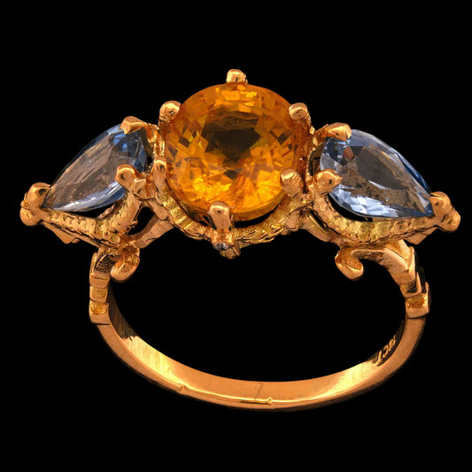 A luxurious 18-carat yellow gold ring featuring a large central yellow sapphire with pear-shaped blue sapphire accents on either side, surrounded by a golden garland setting with four brilliant-cut diamonds on the band.