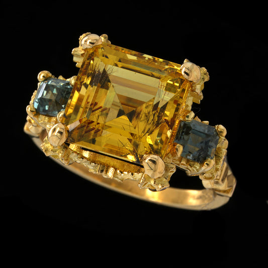 A yellow gold 18k ring featuring a large yellow aquamarine center stone with two smaller teal sapphires on either side, set against a solid black background.