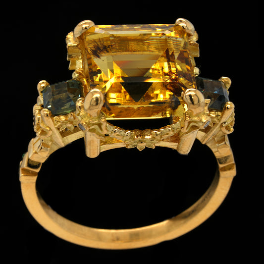 A yellow gold ring featuring a large  yellow aquamarine or heliodor center stone with two smallerteal sapphires on either side, set against a solid black background.
