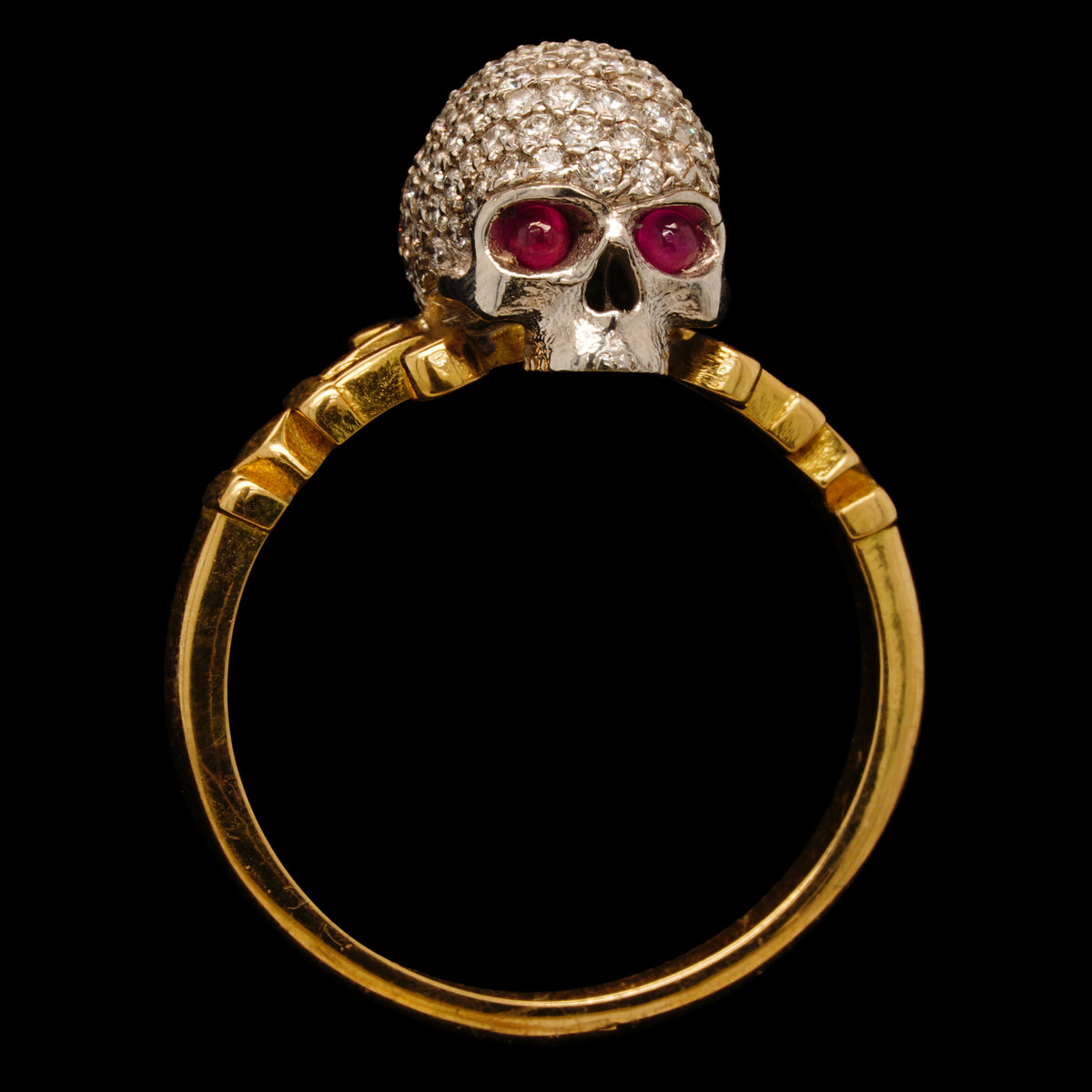An 18kt yellow and white gold ring with a diamond encrusted skull design, featuring piercing ruby eyes, set on a black background.