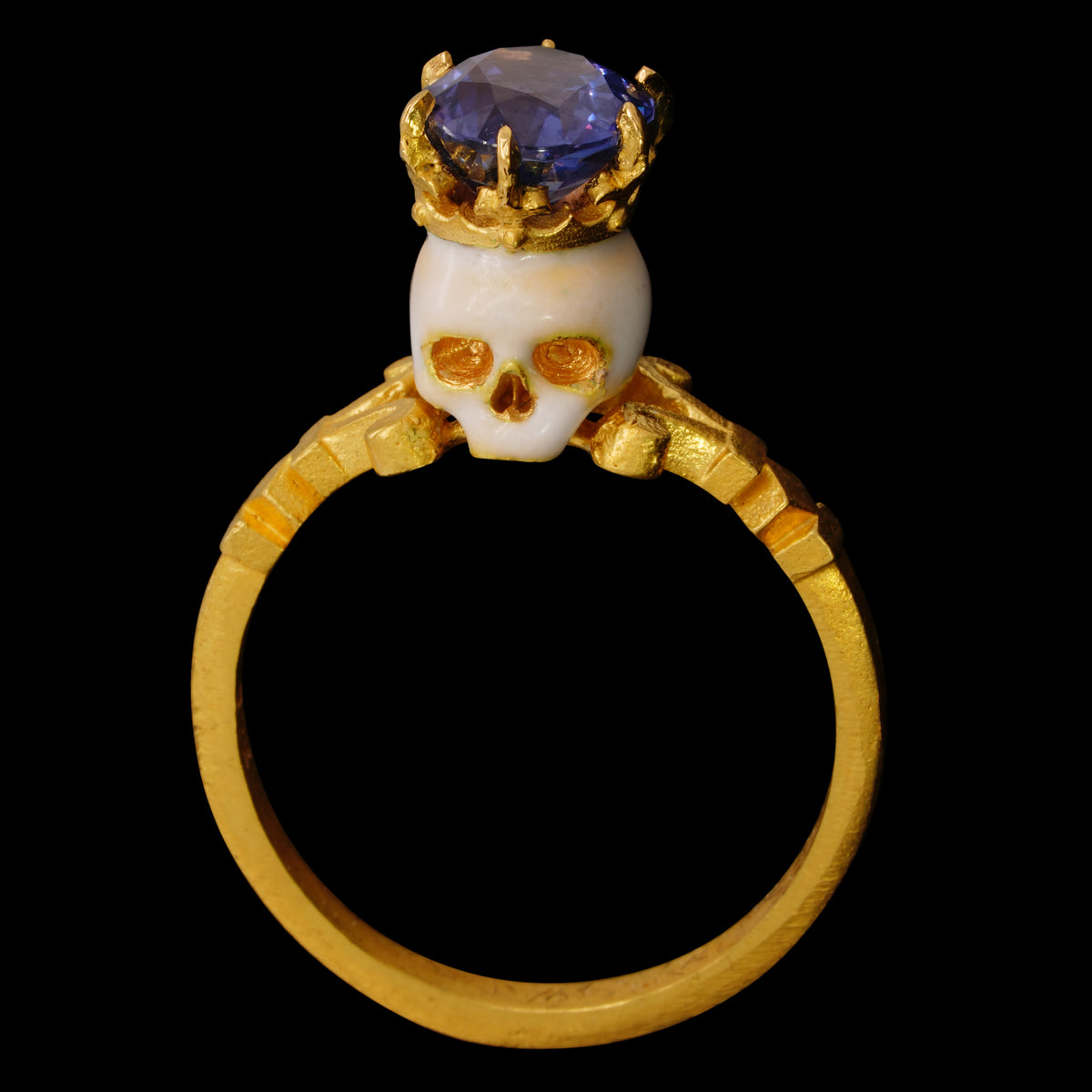A 22kt yellow gold ring with an enamelled skull topped by a crown and a large oval Burmese sapphire with color shift from violet to blue and purple, on a black background.