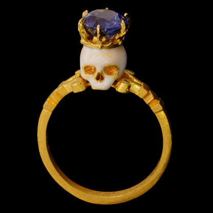 A 22kt yellow gold ring with an enamelled skull topped by a crown and a large oval Burmese sapphire with color shift from violet to blue and purple, on a black background.