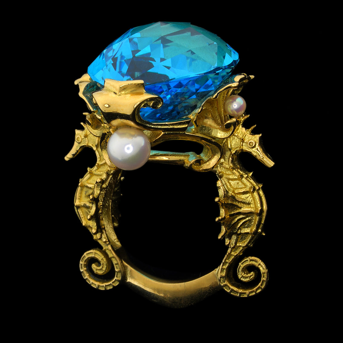 A luxurious 18kt yellow gold ring featuring a large Swiss blue marquise checkerboard cut topaz, surrounded by golden seashells and flanked by two seahorses, with four Akoya pearls. The ring is displayed on a black background.