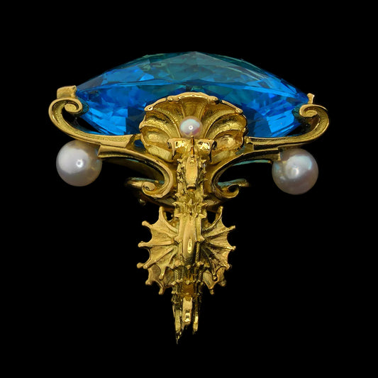 An ornate gold ring featuring a large faceted blue topaz, two white pearls, and a detailed gold scrollwork and seahorses, shown against a solid black background.